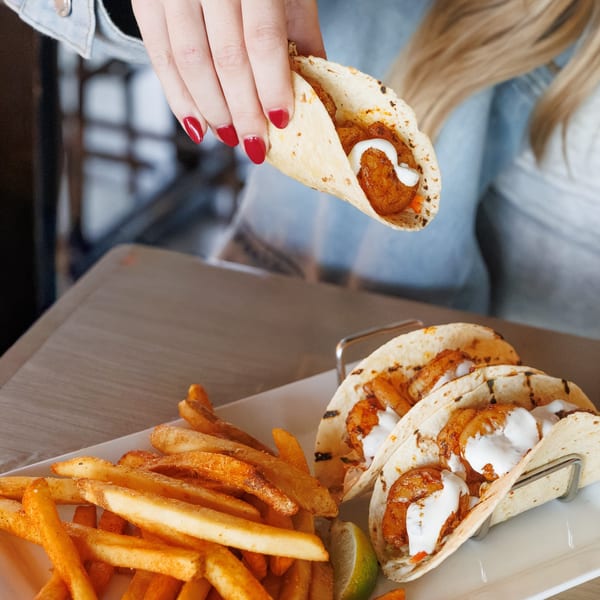 A hand holding a shrimp taco with white sauce over a plate of fries and two more tacos.