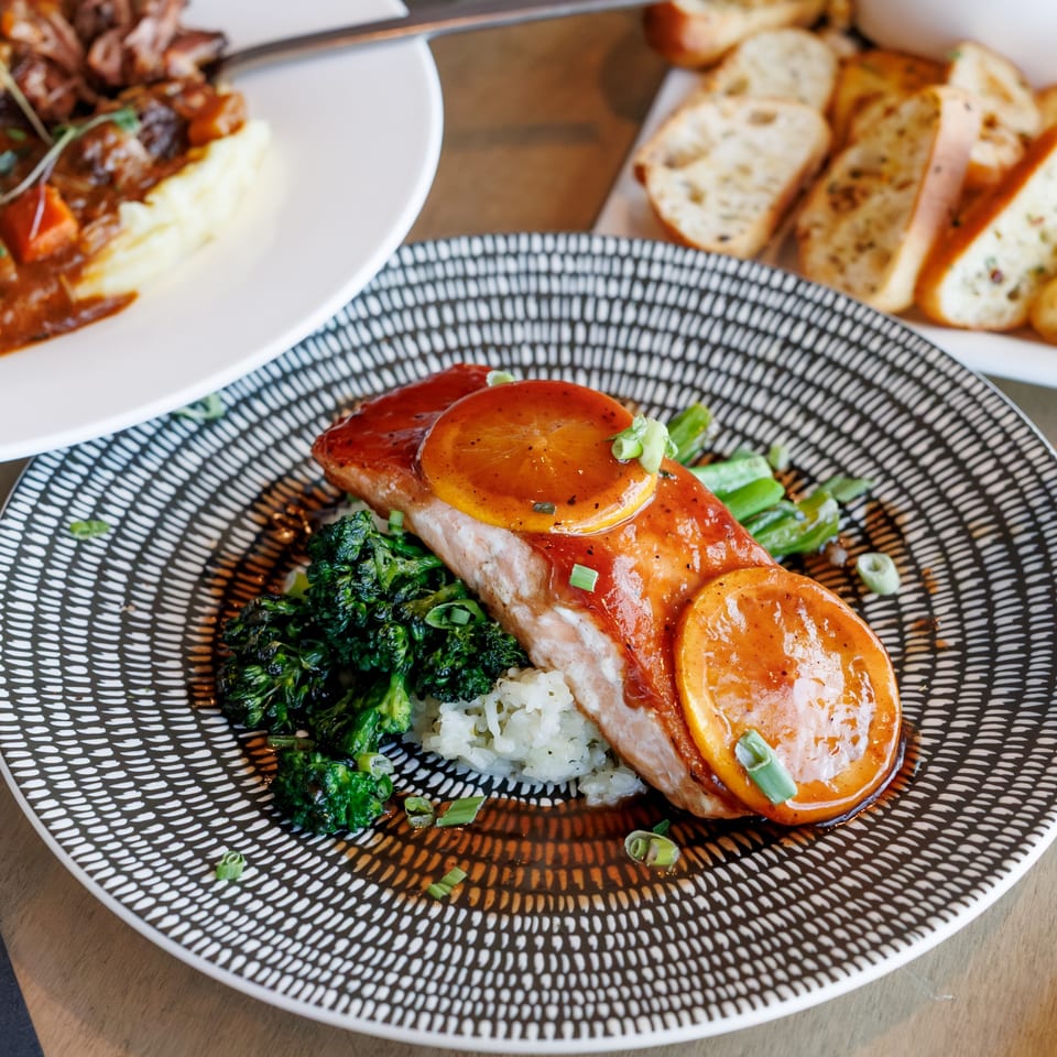 Salmon with orange slices, rice, and broccoli on a patterned plate, with another dish blurred in the background.