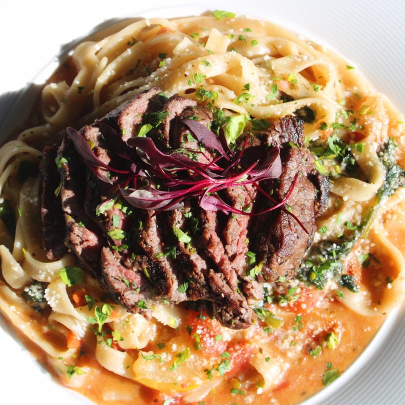 Sliced steak on pasta with a creamy tomato sauce, garnished with fresh herbs and red microgreens.
