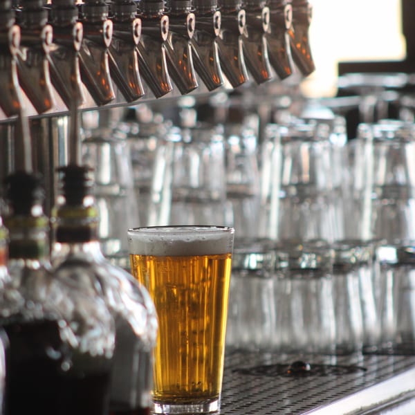 A pint of golden beer with foam sits on a bar counter with beer taps and glasses.