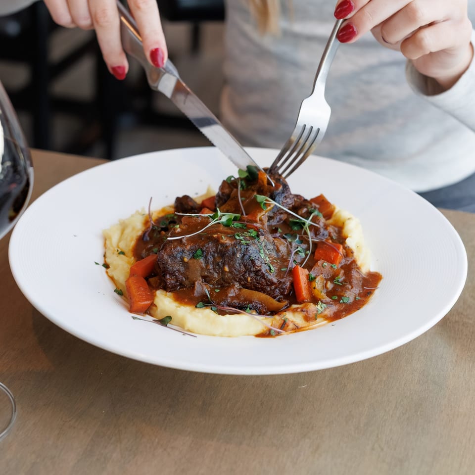 Person cutting into a braised short rib dish with mashed potatoes and vegetables.