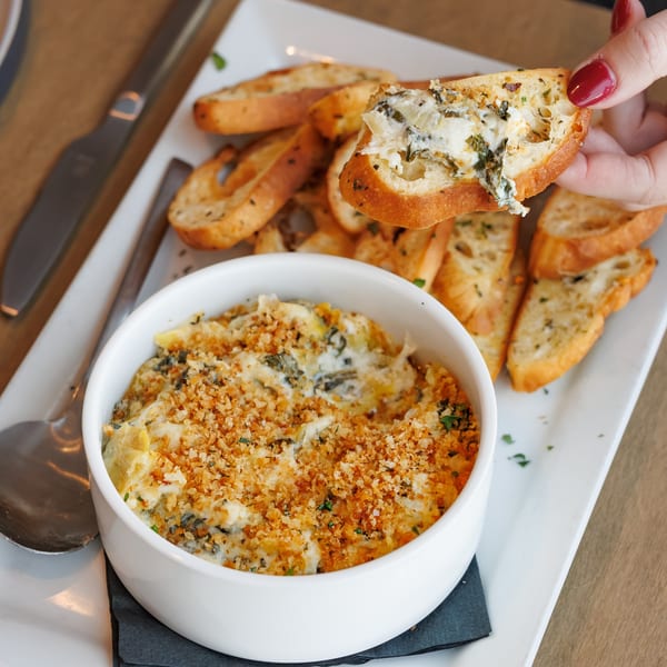 A hand holding a piece of toasted bread with creamy dip, next to a bowl of dip and more bread.