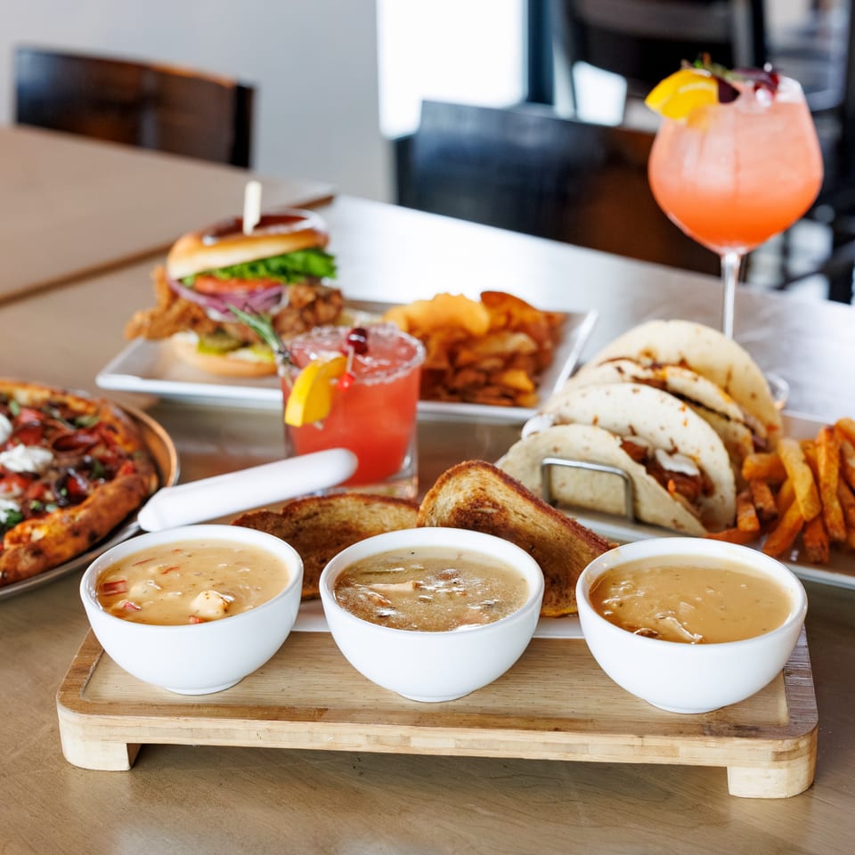 A table spread with various dishes including pizza, tacos, a burger, fries, and soup bowls.