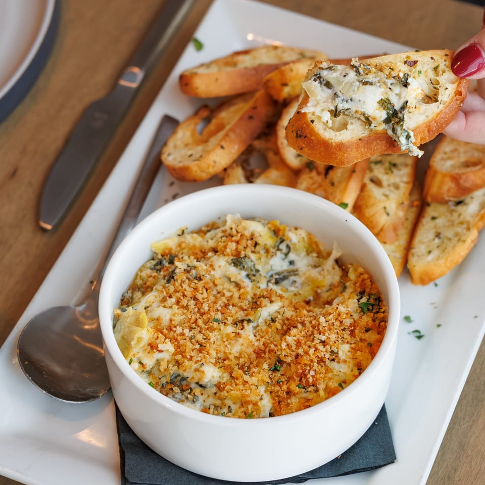 A hand dips a toasted baguette into a bowl of creamy, baked spinach and artichoke dip.