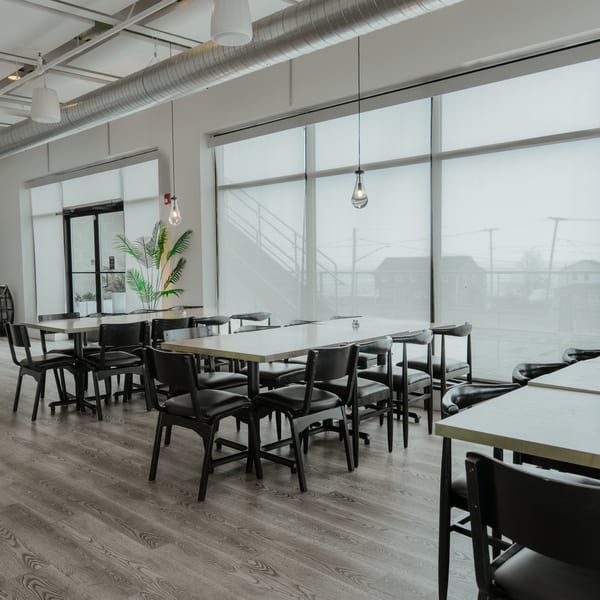 Modern restaurant dining room with light wood tables, black chairs, large windows, and exposed ductwork.