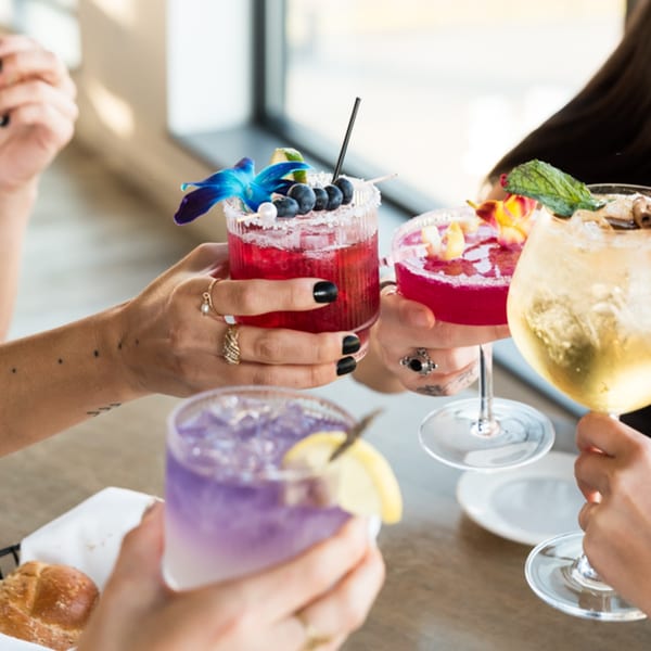 Four hands holding colorful cocktails with various garnishes, clinking glasses in a bright setting.