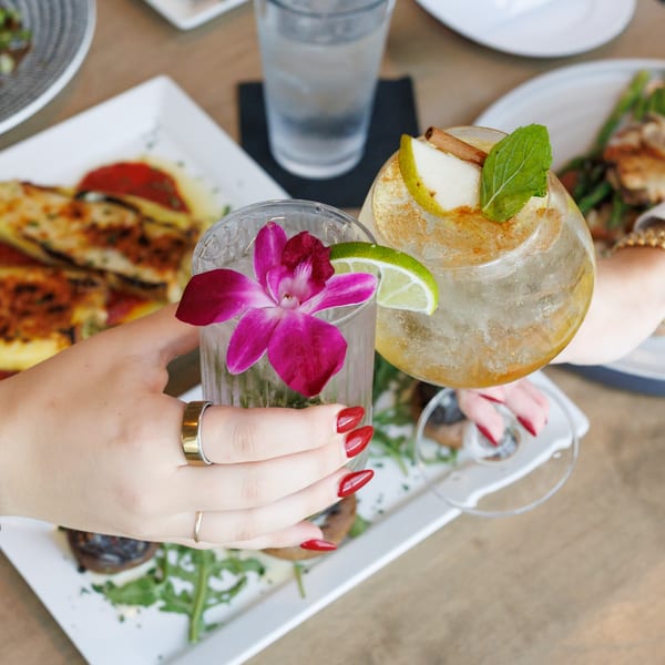 Two hands clinking cocktails, one garnished with a pink orchid, the other with pear and mint.
